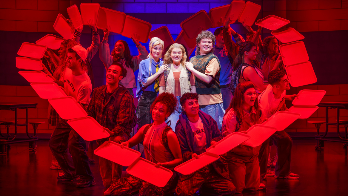 Three young people smile as they are framed by classmates holding red lunch trays in the shape of a heart