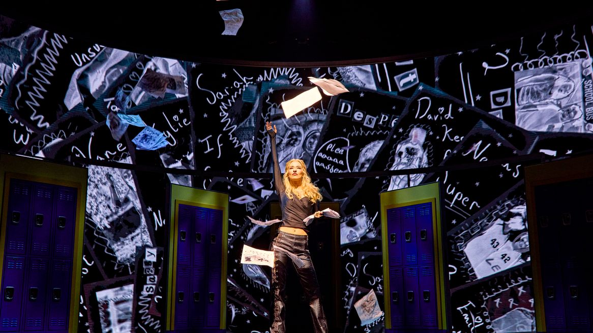 A girl in all black throws papers into the air in a school hallway