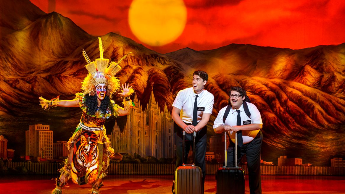 Two young Mormon men stand with suitcases looking excited while a woman in African dress sings