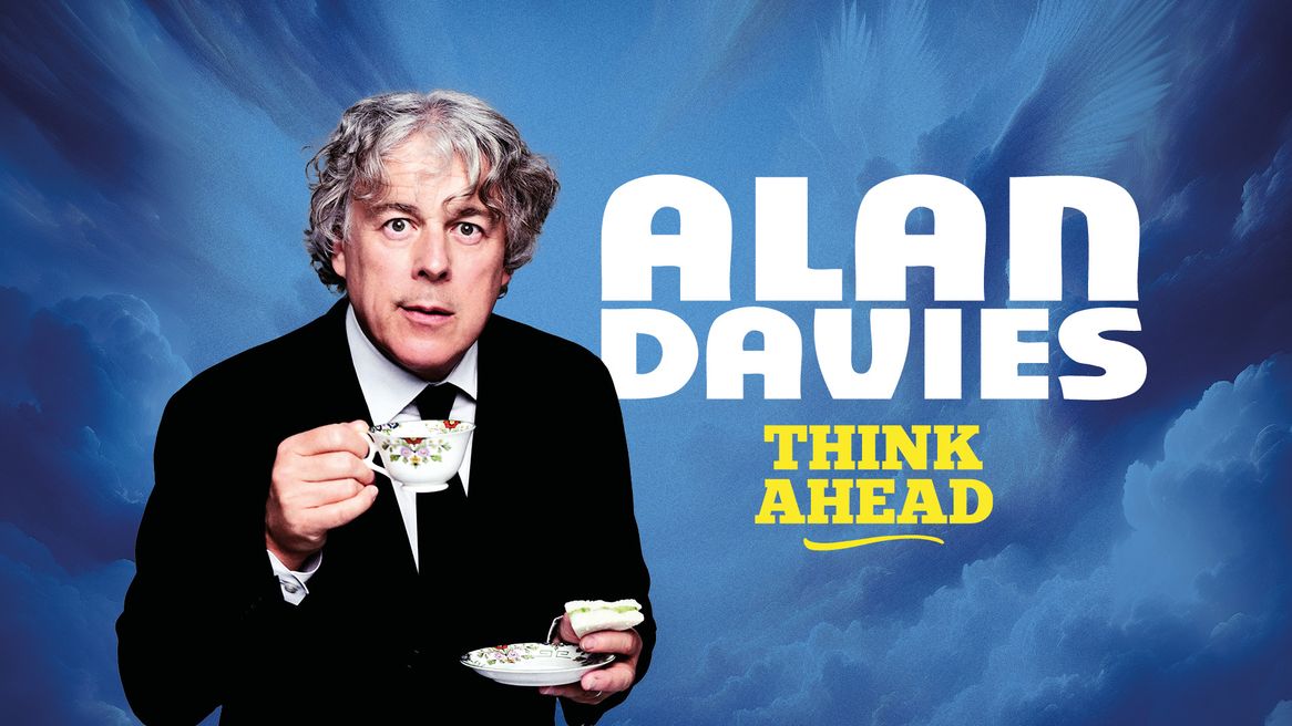 Comedian Alan Davies in a black suit, whit shirt, black tie lifting a china style cup with a blue sky cloudy background