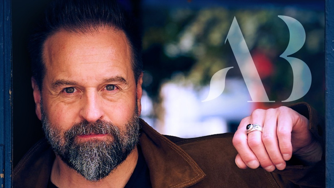Alfie Boe in brown jacket with dark v neck t-shirt leans against a blue wooden door frame looking straight at the camera.