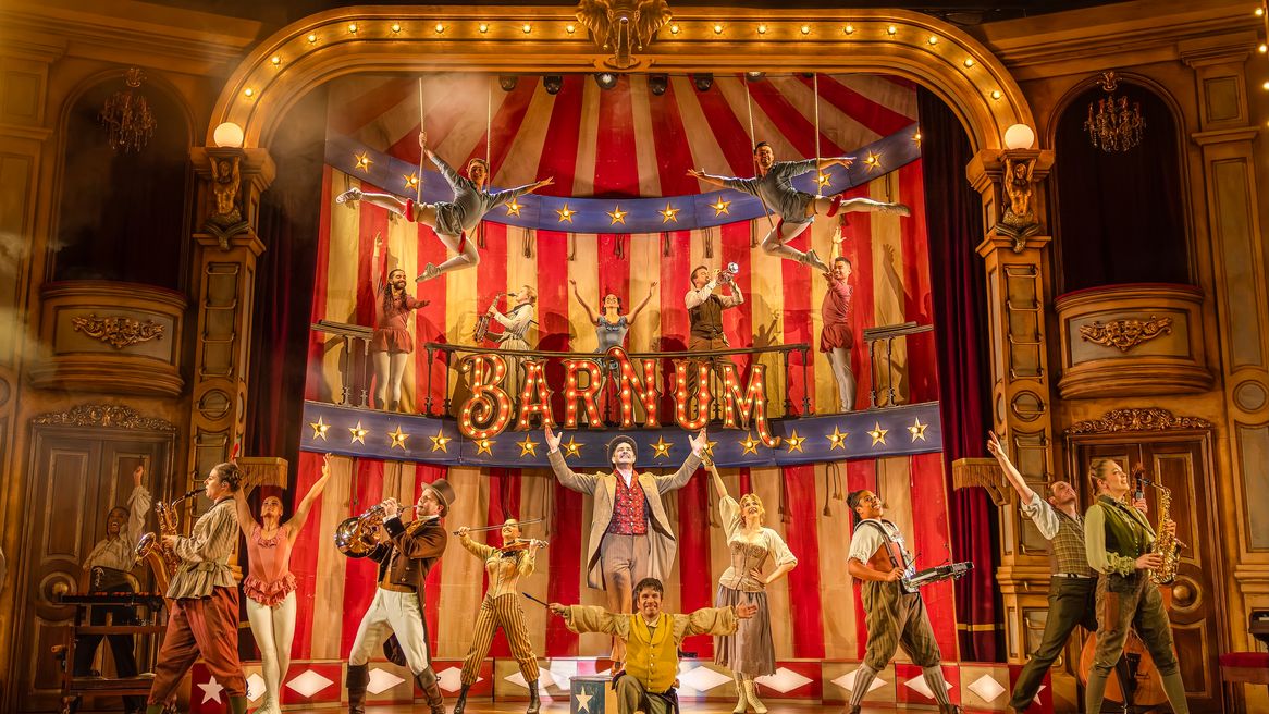 A group of performers in circus outfits dance and play instruments in a big top