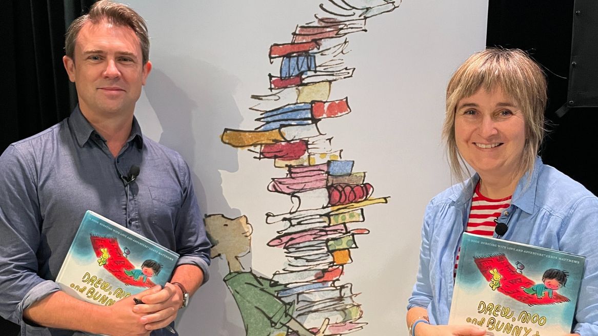 A man and woman in blue open necked shirts clutch copies of the book Drew, Moo and Bunny Too