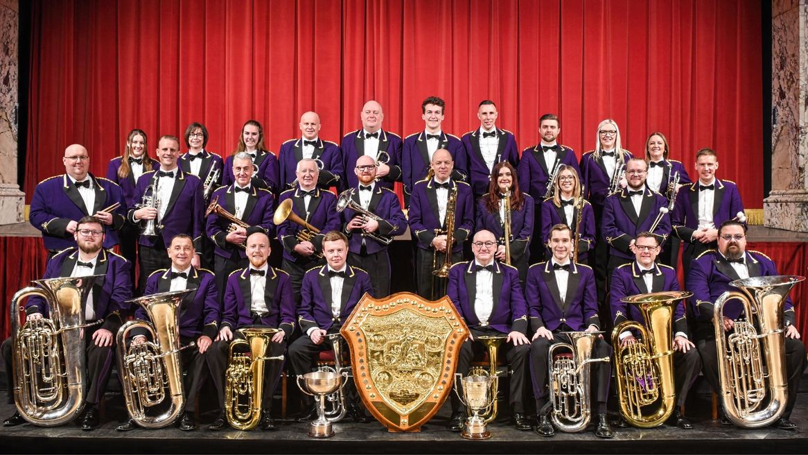 A mixed group of male and female musicians wearing purple jackets, white shirts and black ties, pose with their instruments