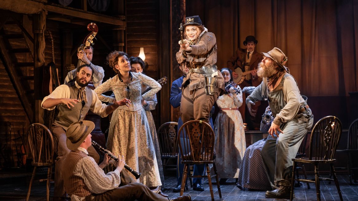 A woman in a Western outfit poses with a gun while stood on a chair as onlookers watch