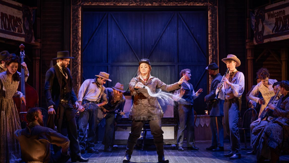 A woman in a Western outfit pours salt onto a stage in front of a band as men look on