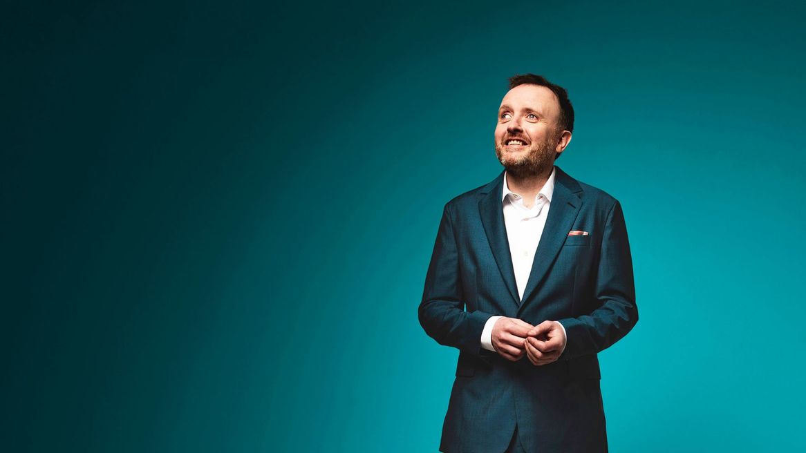 Comedian Chris McCausland in a blue suit looking up to the sky with a blue background. 