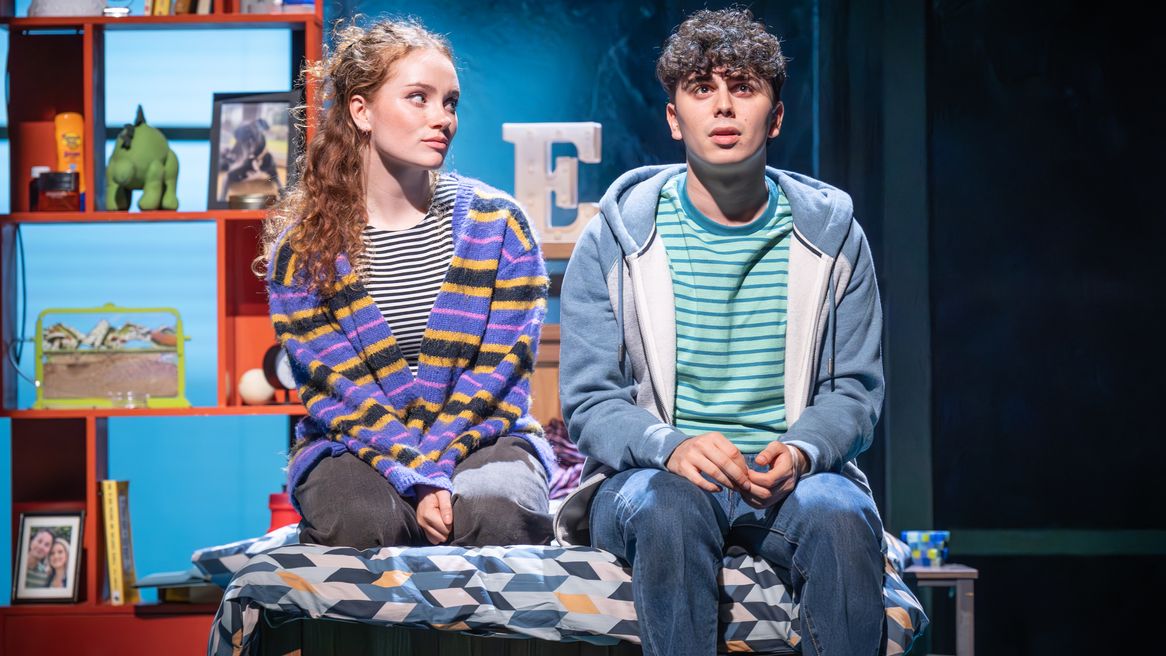 A teen girl and a teen boy sit on a bed in the boy's bedroom