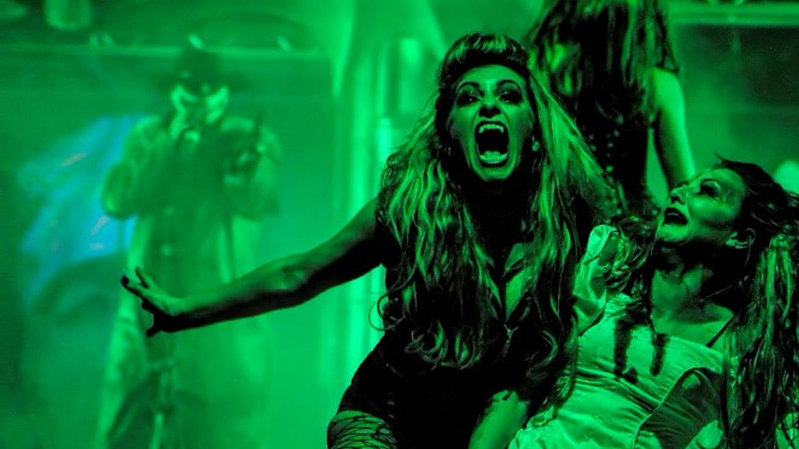 A female vampire bears her fangs after biting another woman who is screaming, the image is awash with green neon. 