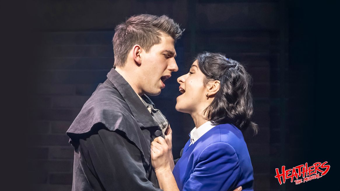 A girl in a blue blazer holds onto a boy wearing a trench coat as they sing