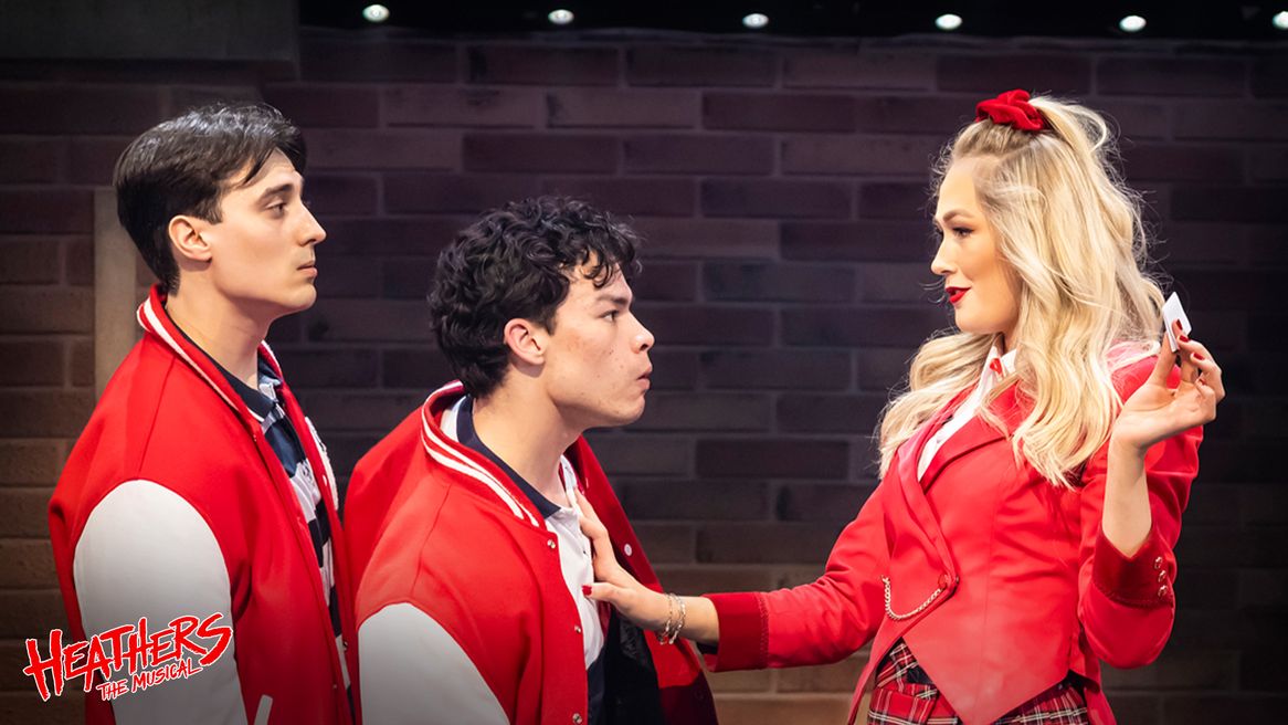 A girl in all red holds up a note in front of two boys in letterman jackets