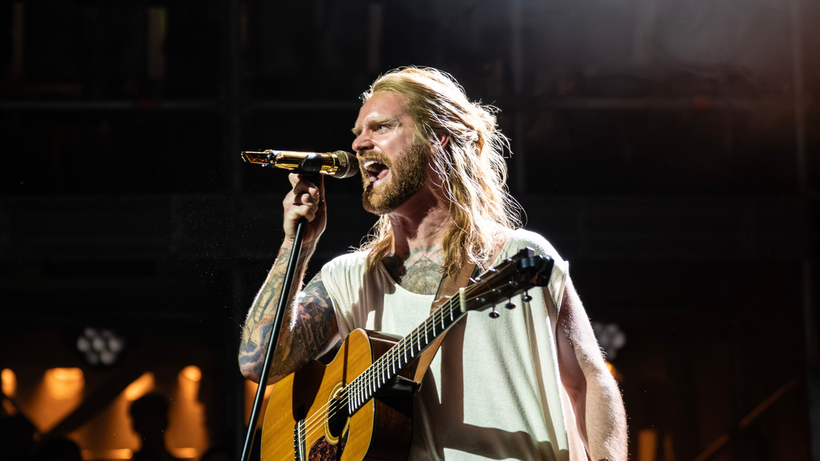 Sam Ryder as Jesus dressed in cream colured vest-style top sings angrily into a microphone with guitar strapped to his chest