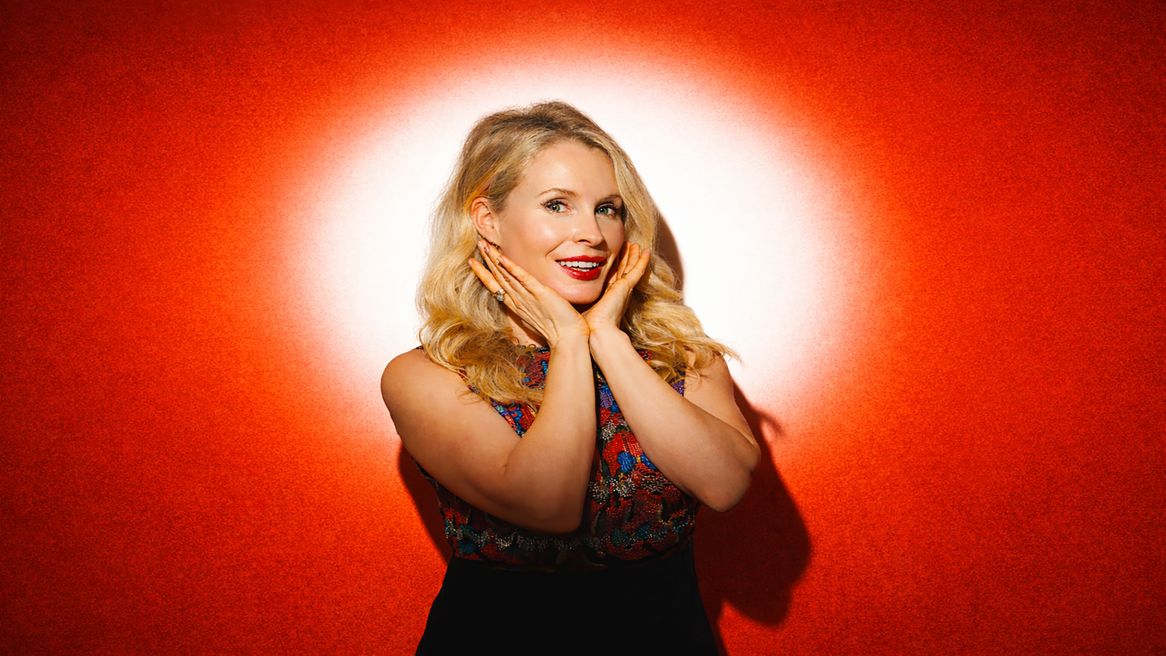 Highlighted by a white spotlight, Lucy Beaumont smiles with her hands cupping her chin in front of an orange backdrop.