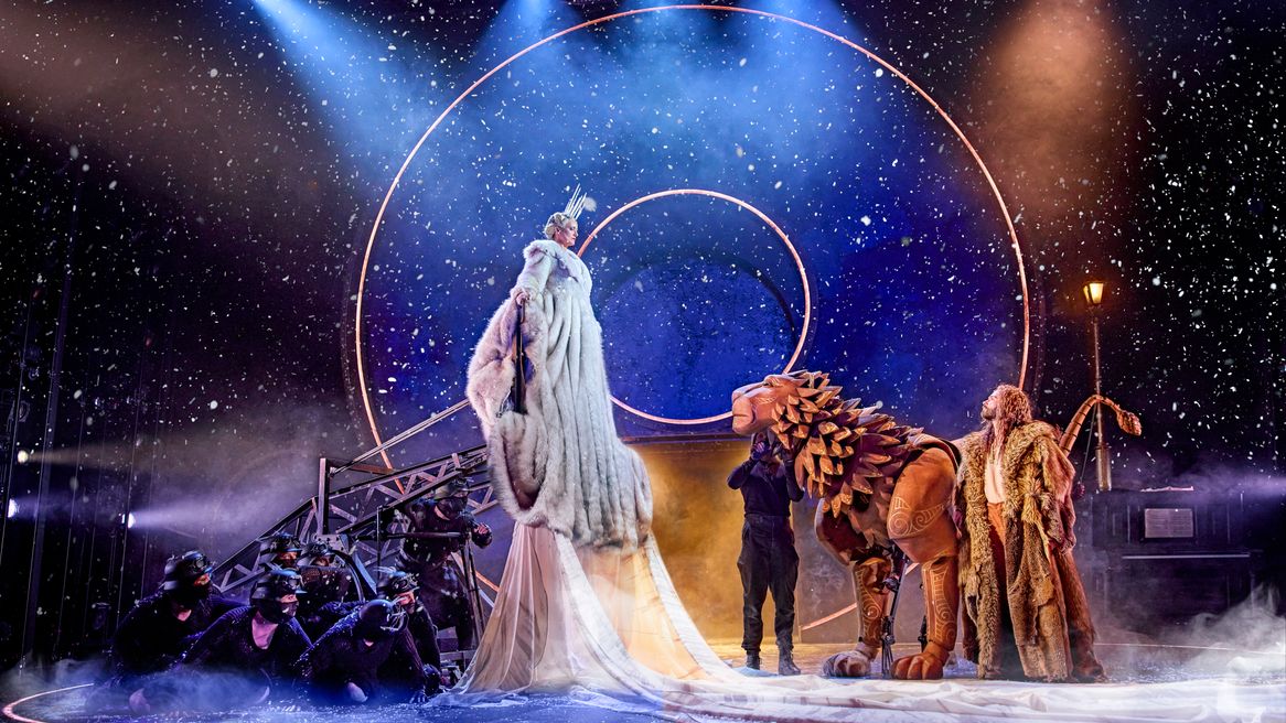 A witch in a fur coat with an icy crown towers over a lion controlled by puppeteers