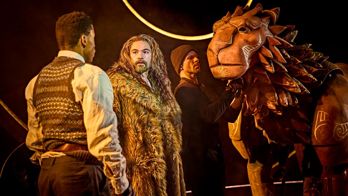 A lion puppet and a man with long hair and a fur coat listen as a young boy speaks