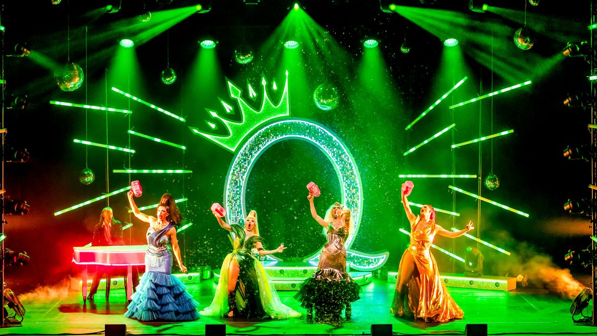 Four drag queens in gowns perform together on a green lit stage