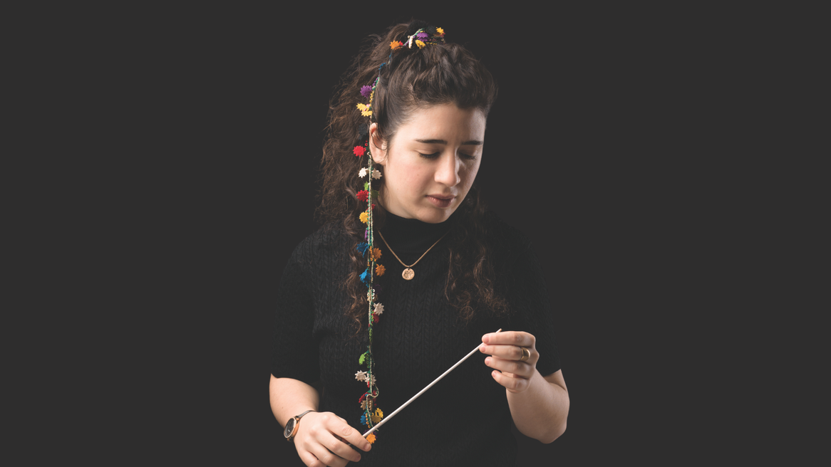 A woman with flowers threaded into her hair looks down holding a conducting baton