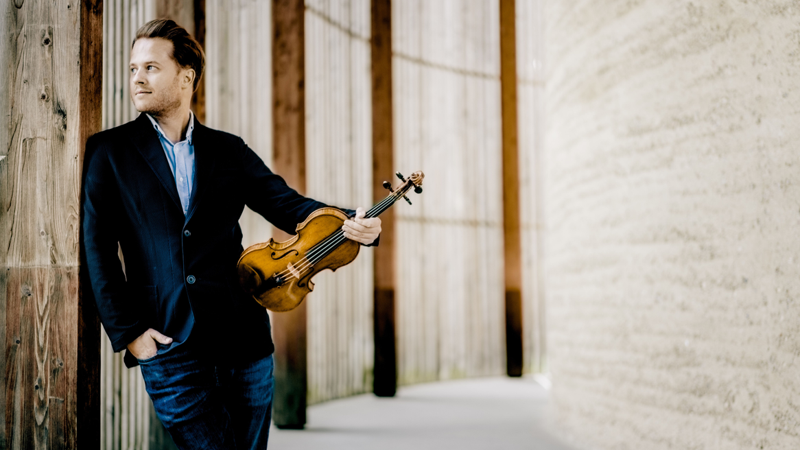 A well-dressed man leans against a wall holding a violin