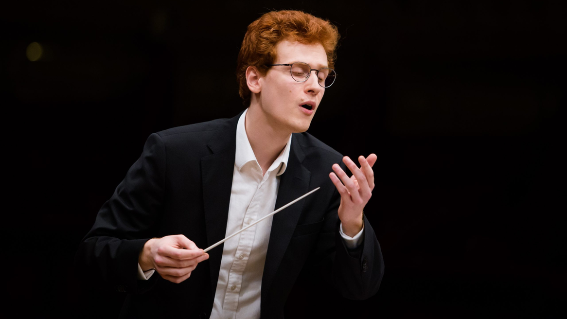 A man in a suit conducts an orchestra with his eyes closed