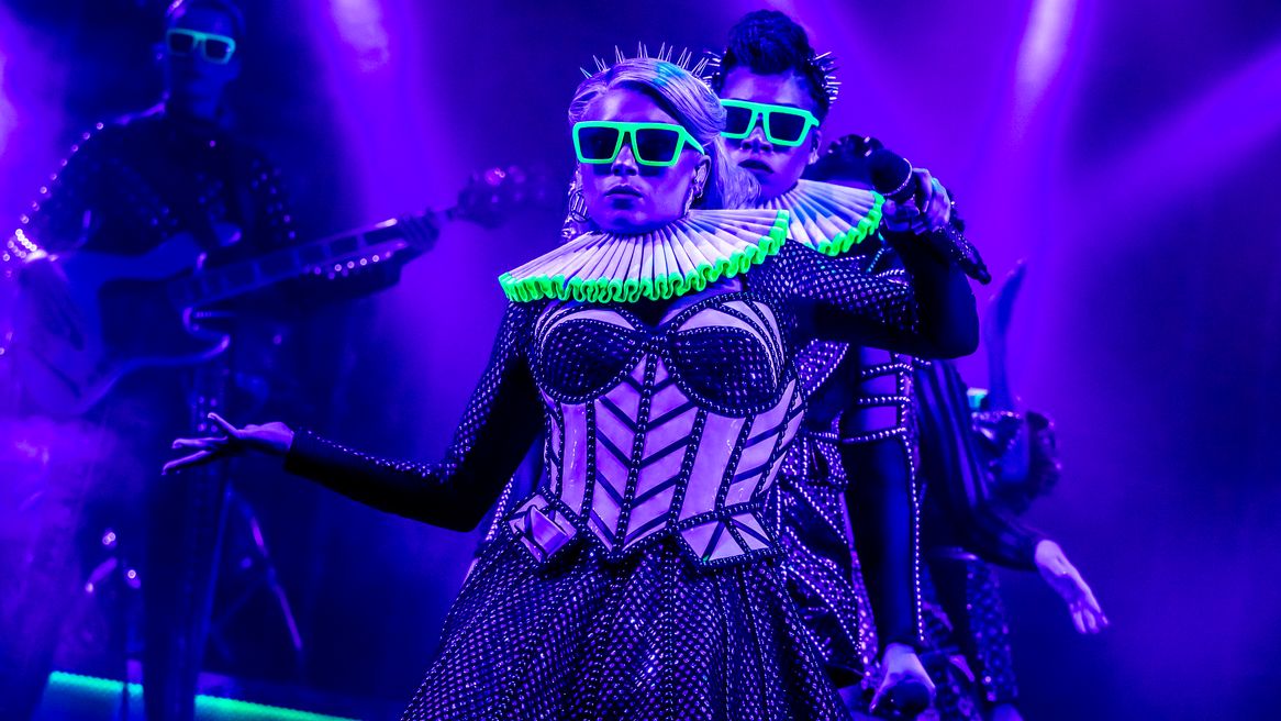 A woman in a Tudor inspired popstar outfit poses with neon sunglasses and a neon ruff