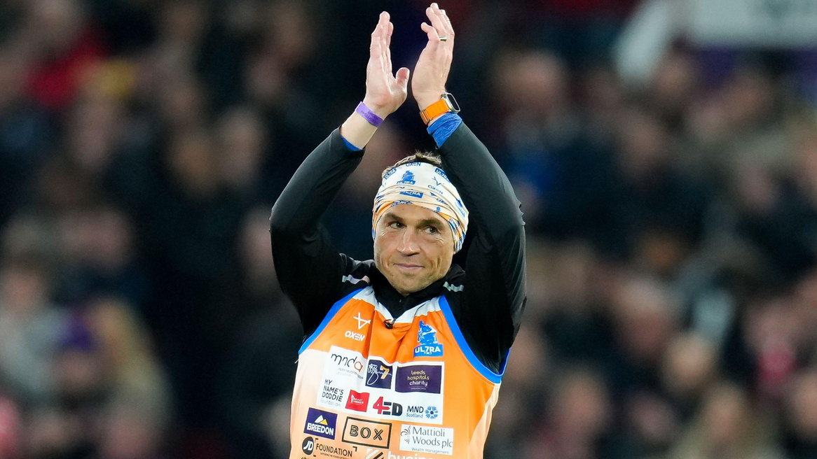 Kevin Sinfield dressed in orange sporting bib with long sleeve black top underneath, claps his hands above his head.