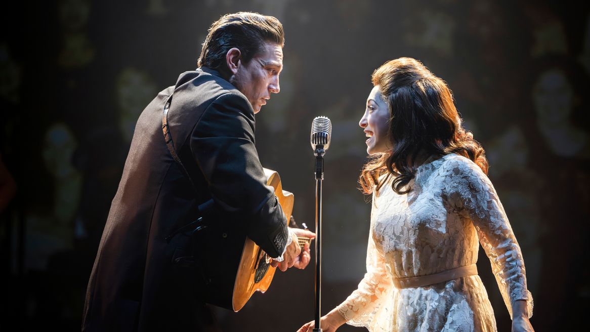 A man playing guitar and a woman in a white dress sing together into an old fashioned microphone