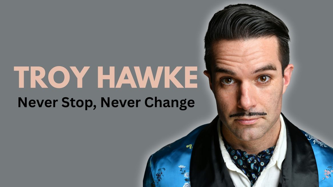 A dark haired, moustached man in a blue satin jacket and cravat stares straight into the camera with a serious expression.