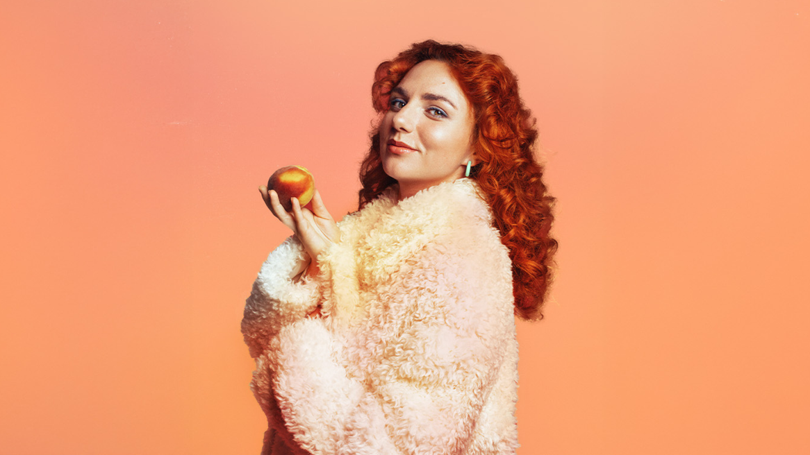 A woman with curly red hair wearing a fuzzy peach coloured coat holds a peach in her hand