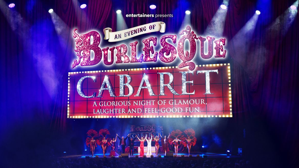 Cabaret performers in red sequin dresses with feather headdresses stand with arms aloft on stage as if taking a bow.
