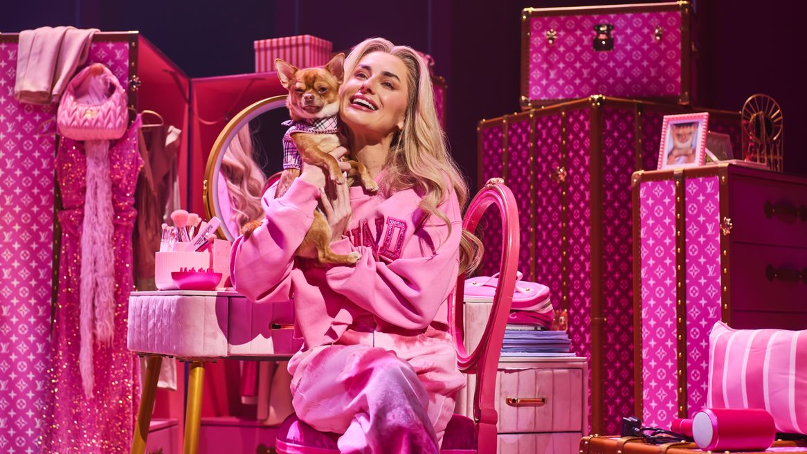 A blonde woman in a pink tracksuit sits in front of a pink dressing table while holding a chihuahua