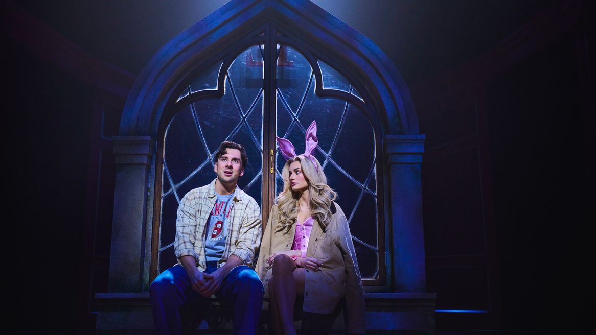 A blonde woman in a pink bunny costume sits in front of a large window with a casually dressed man