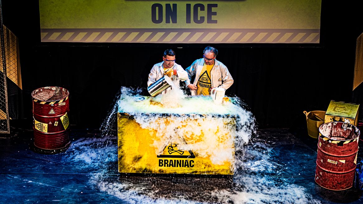 Two men dressed in white lab coats pour liquid into a lagre square tub with a toxic sign on the front