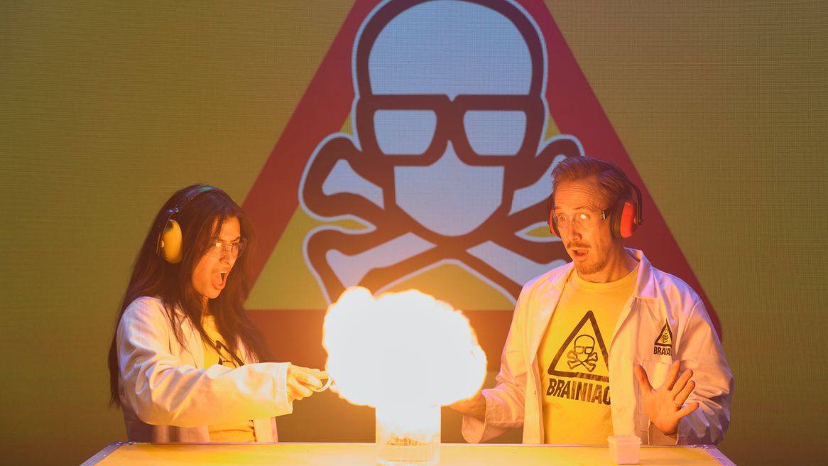 A Man and a woman in white coats stand in from of a skull and cross bones sett fire to a liquid in a glass jar