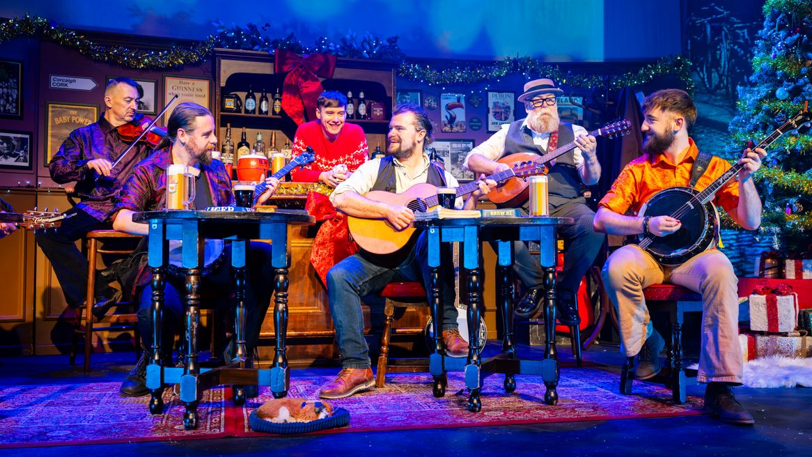 Six performers with instruments sit in a pub decorated for Christmas with a little dog in a bed on the floor