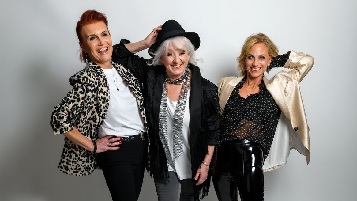 Three women laugh and pose together in black, white and cream outfits