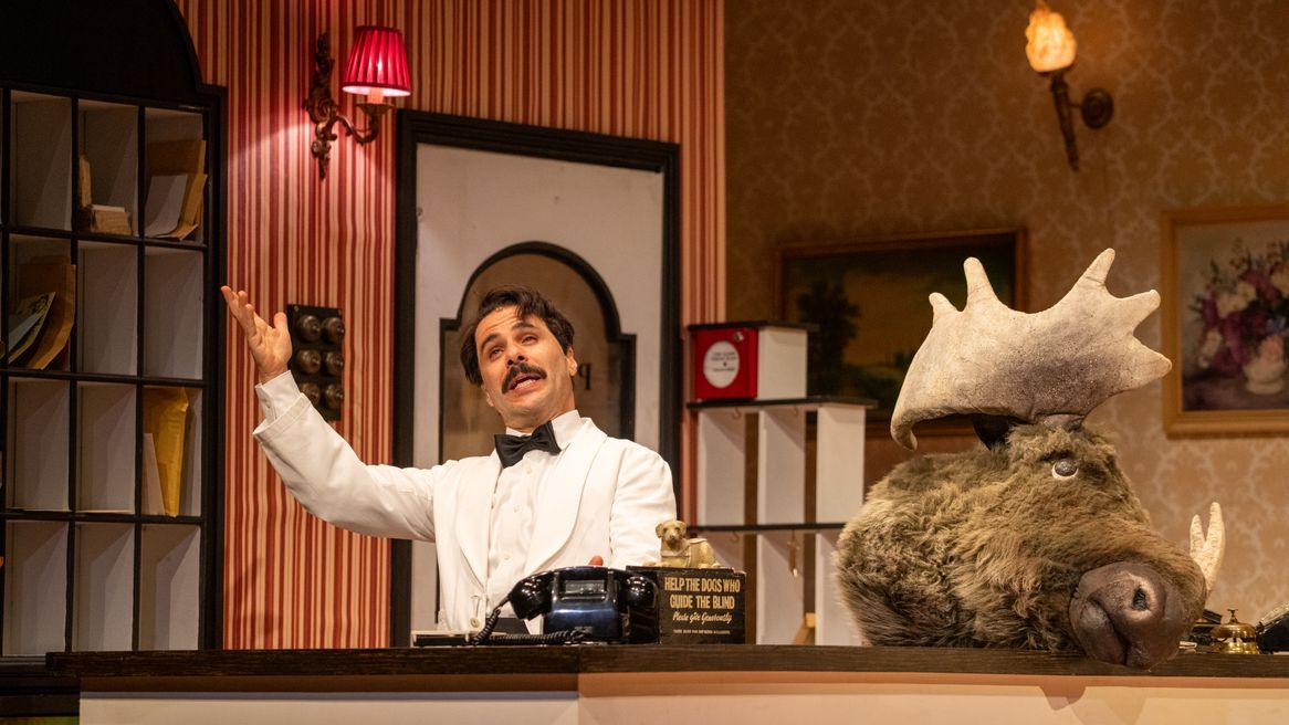 A man in a white suit with black bow tie waves his arm next to a large moose head