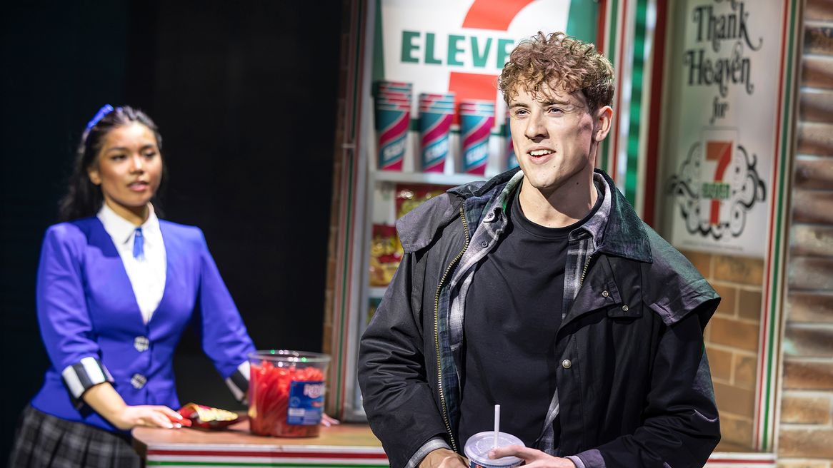 A girl in a blue blazer looks on as a boy in a trench coat sings in a 7/11 convenience store