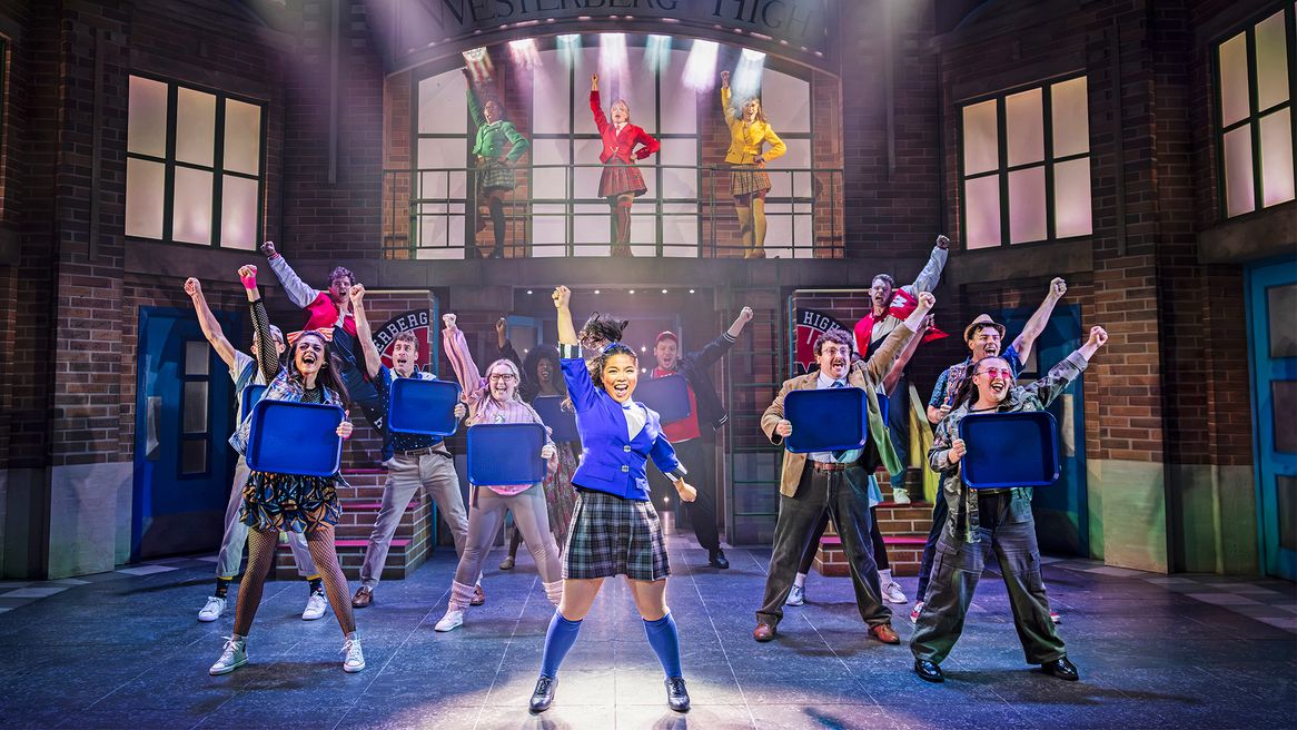 People stand with their fists raised, holding school lunch trays as three girls stand on a balcony with their fists raised