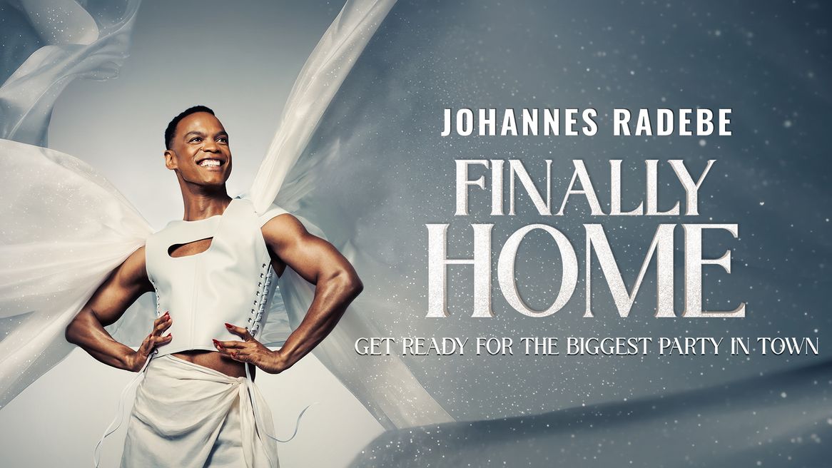 Johannes Radebe smiles wearing an all white outfit with a long white glittery cape