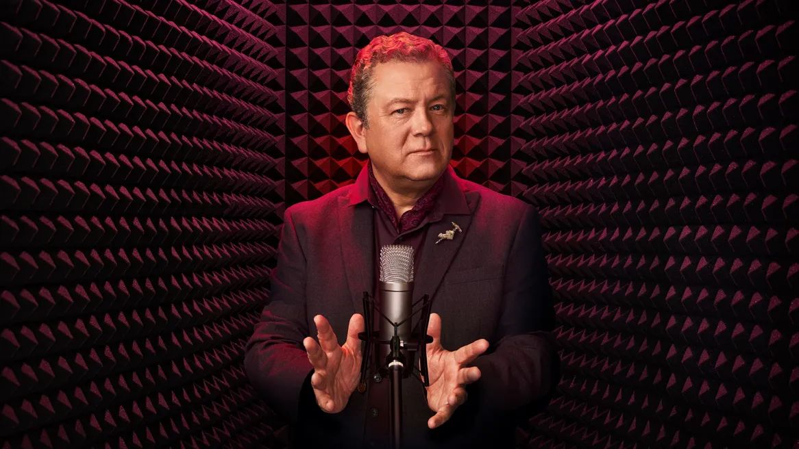 Comedian Jon Culshaw in a purple suit in front of a microphone with a purple background