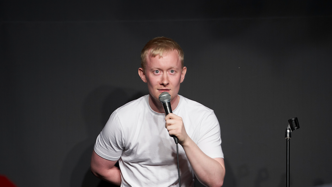 Comedian in a white T-shirt and bleached blonde hair holding a microphone staring at the camera