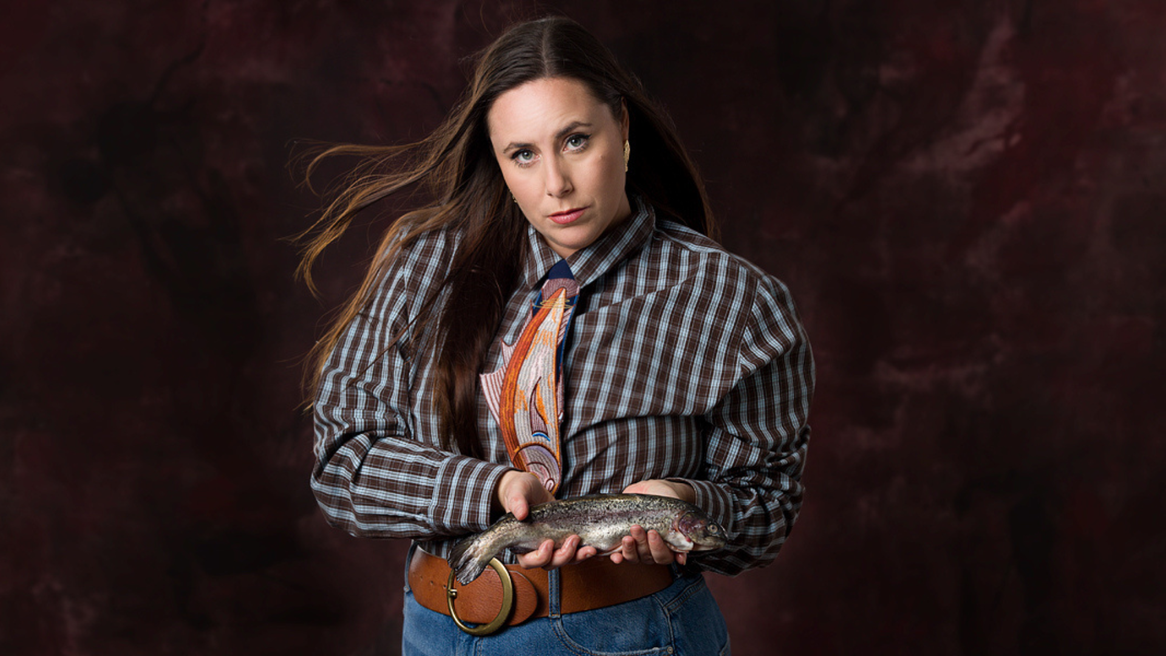 A woman wearing a fish shaped tie holds a trout