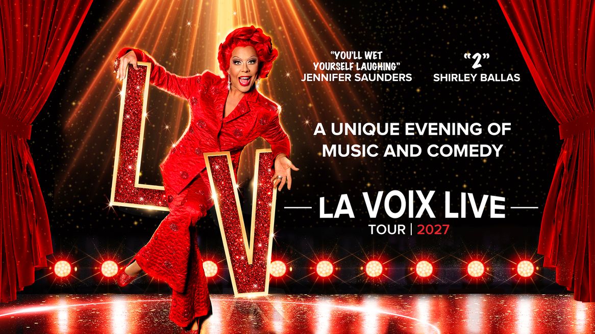 A drag queen in a short redwig and a red suit poses on a glittery red L and V on stage