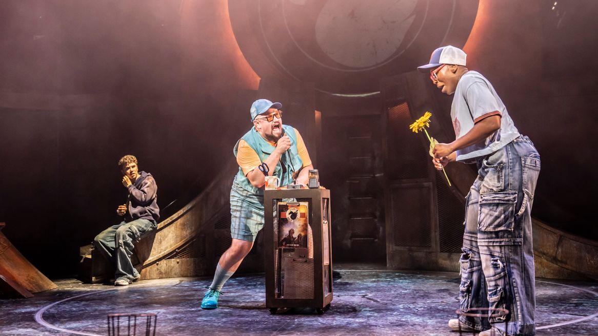 Three characters on stage one holds a flower while another leans over a box and a boy sits down in the background