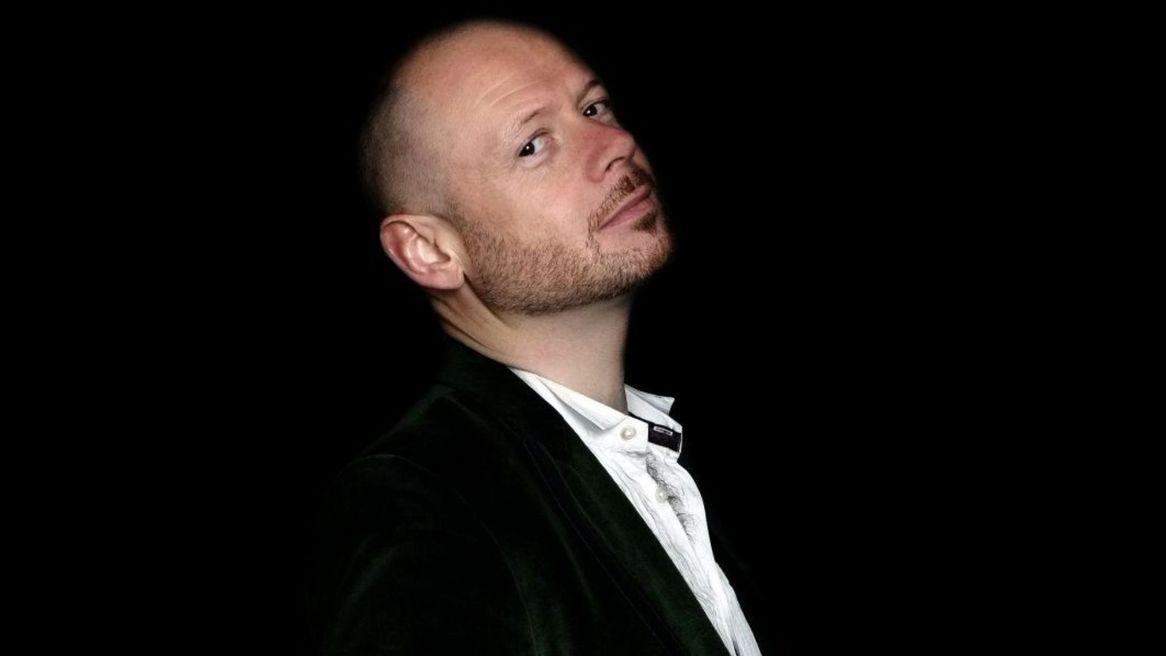 Comic Alistair Barrie in a black suit looks over his shoulder