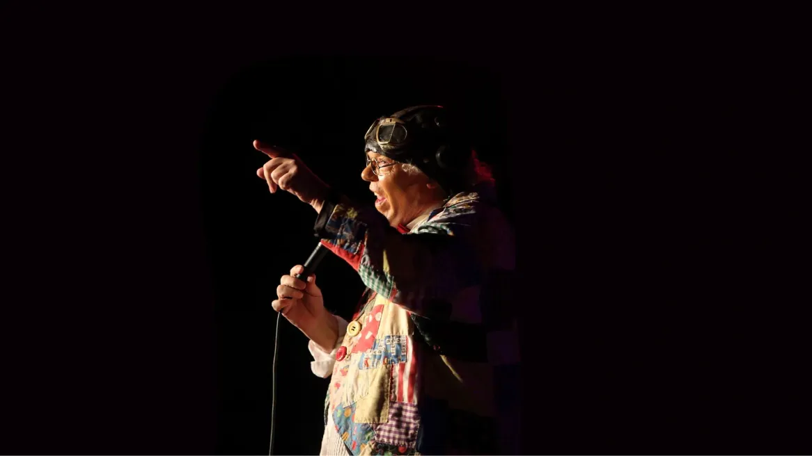 A man in patchwork jacked with leather flying cap and goggles on his head points at the audience as he talks into microphone