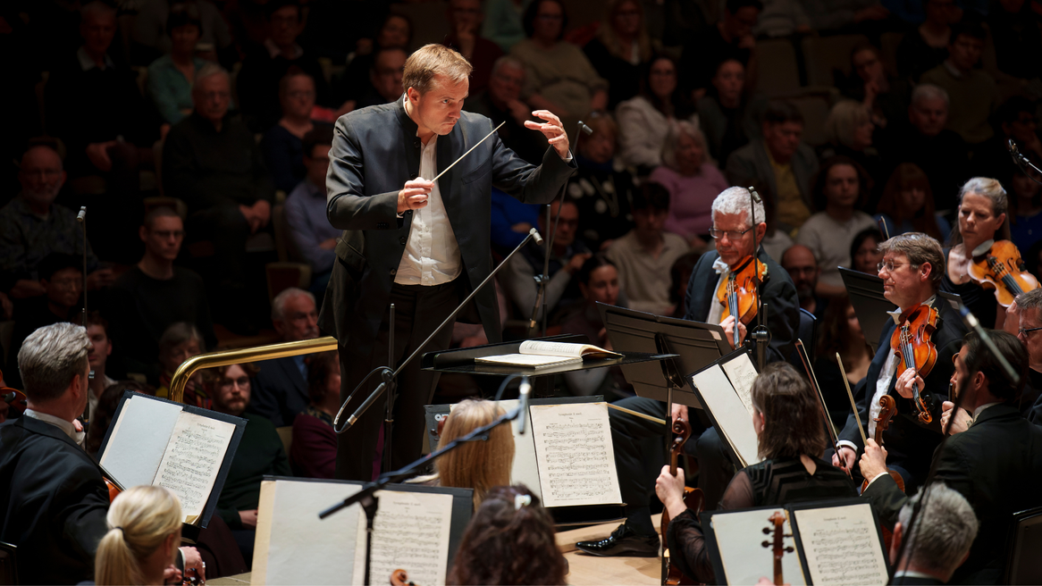 A male conductor faces an orchestra with one armed raised holding a baton and a face of concentration