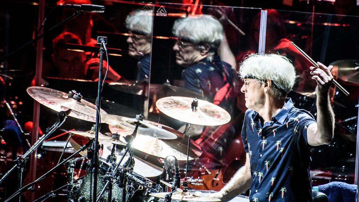 Man sat at a drum kit with one arm raised with a drum stick ready to hit the drums