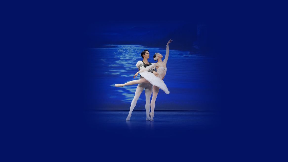 A male ballet dancer holds a female ballet dancer around the waist in front of a lake