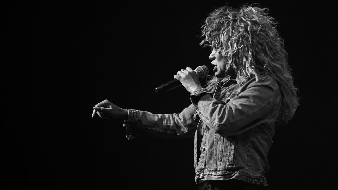 A woman in a Tina Turner inspired wig sings into a microphone